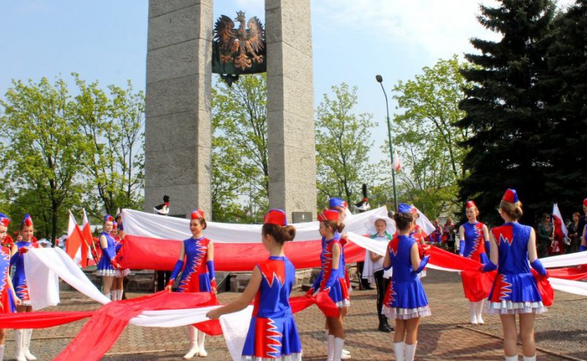 Kilkanaście dziewczyn w niebieskich strojach trzyma szarfy w barwach flagi Polski, za nimi pomnik z metalowym godłem
