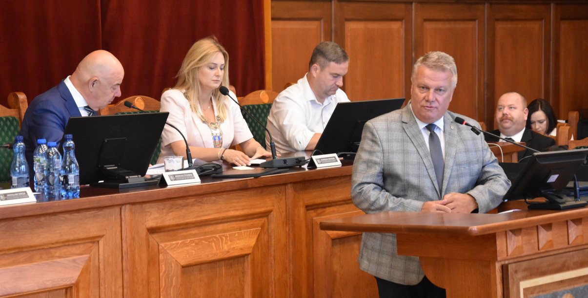 Burmistrz Dzierżoniowa przy mównicy, za nim przy stole prezydialnym przewodnicząca i dwóch wiceprzewodniczących rady