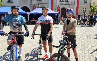 Trzech rowerzystów na rynku, za nimi niebieskie namioty
