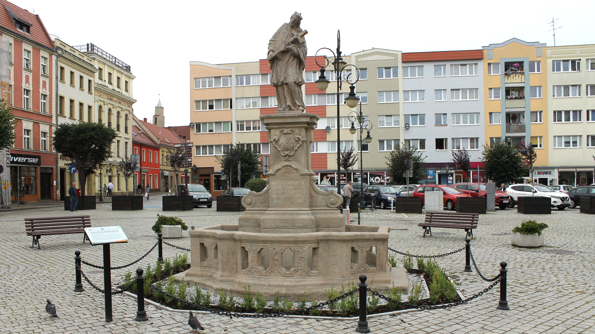 Rzeźba Jana Nepomucena na cokole, otoczaona kamiennymi barierkami, w tle budynki