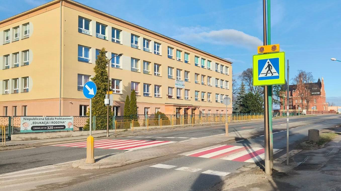Szeroka droga, biało-czerwone przejście dla pieszych, za drogą duży trzypiętrowy budynek