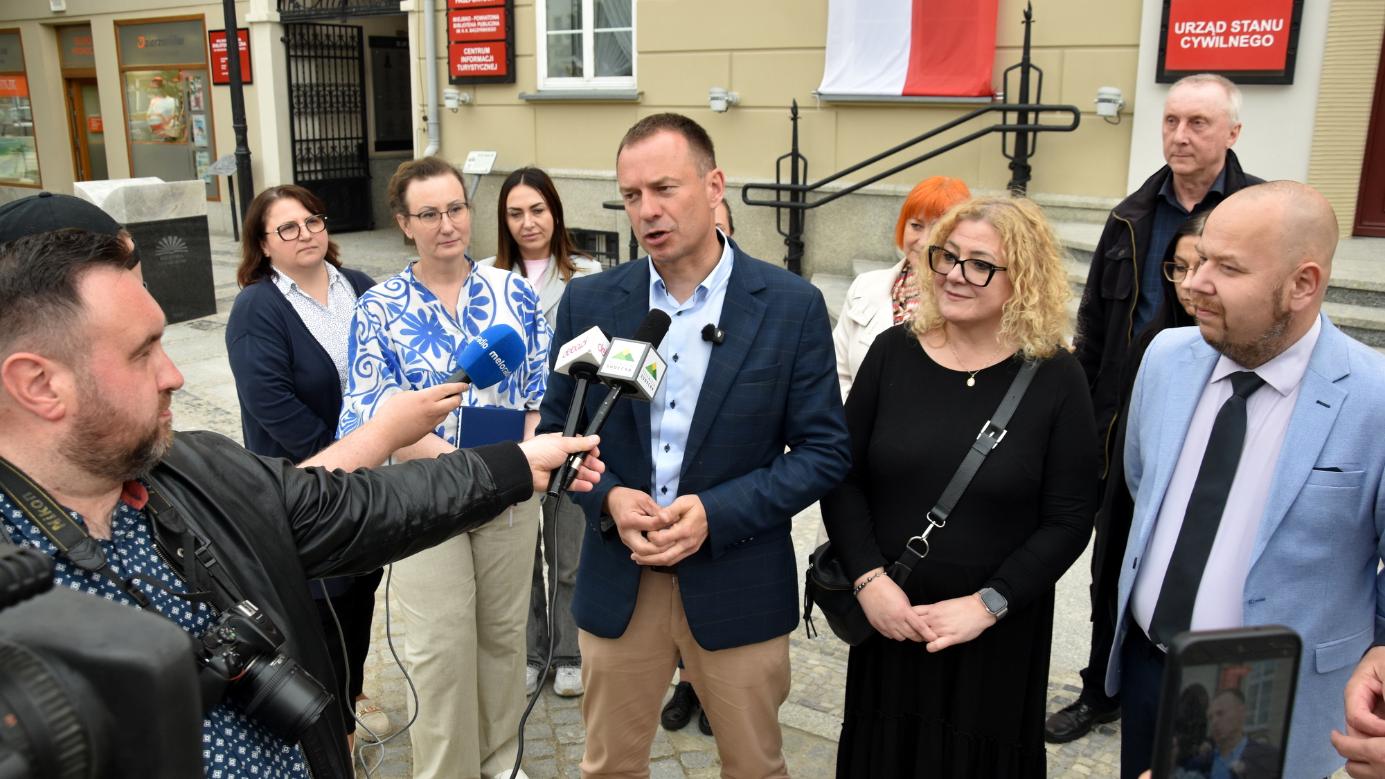 wiceminister mówiący do mikrofonów, dookoła niego grupka ludzi