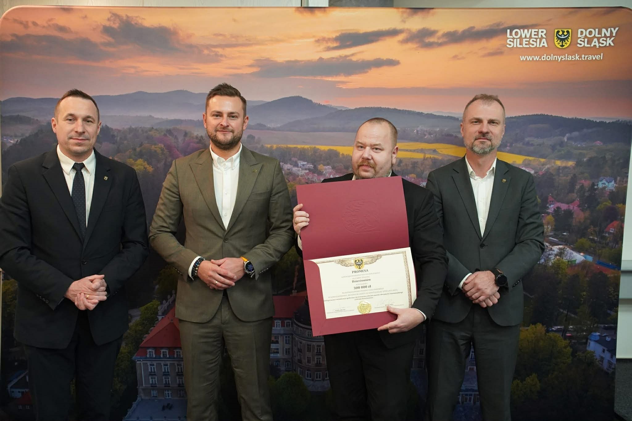 Czterech mężczyzn na tle ścianki promocyjnej województwa dolnośląskiego, zastępca burmistrza Radosław Michałek (drugi od prawej) trzyma promesę