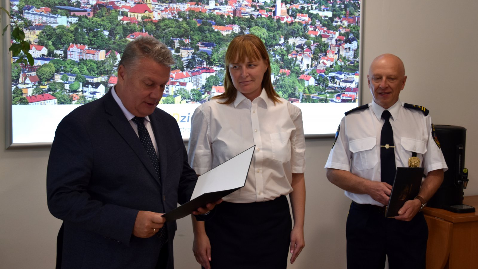 Mężczyzna w garniturze odczytuje list, po jego prawej strażniczka i strażnik miejski