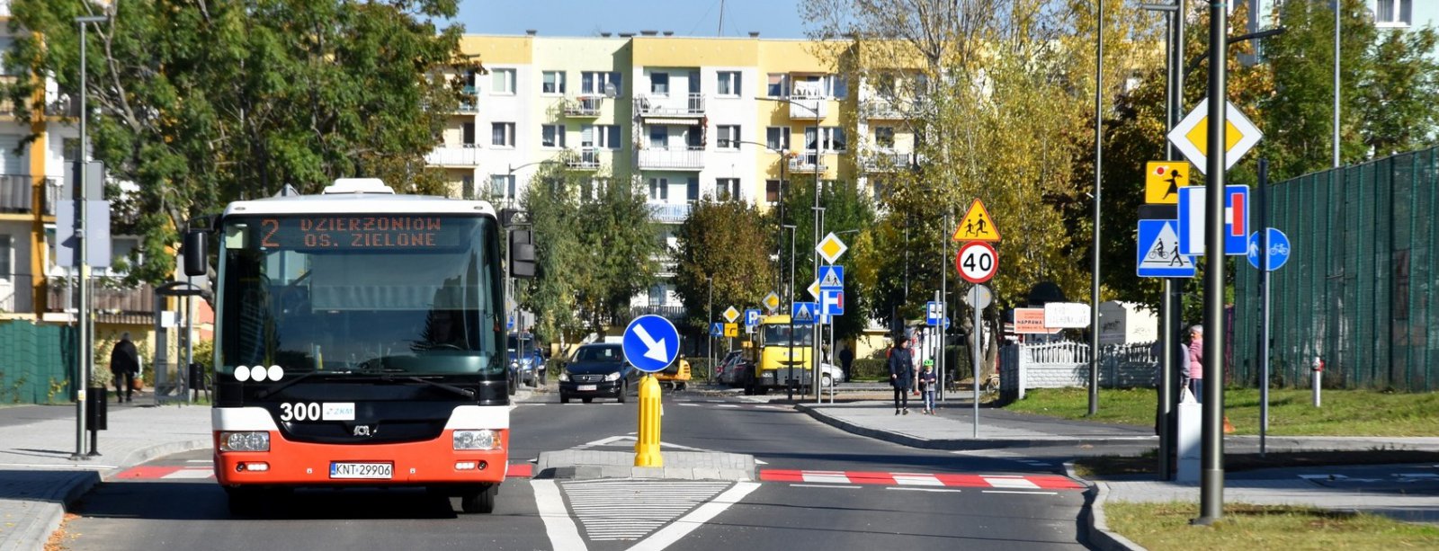 Autobus komunikacji miejskiej na osiedlowej drodze, przed nim czerwono-białe przejście dla pieszych