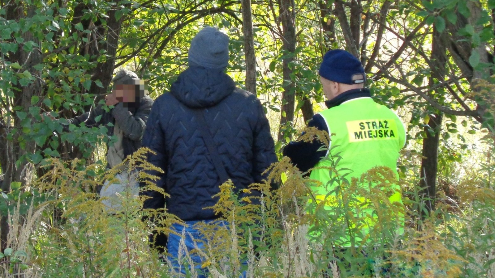 Strażnik miejski w jaskrawej kamizelce, obok dwie osoby