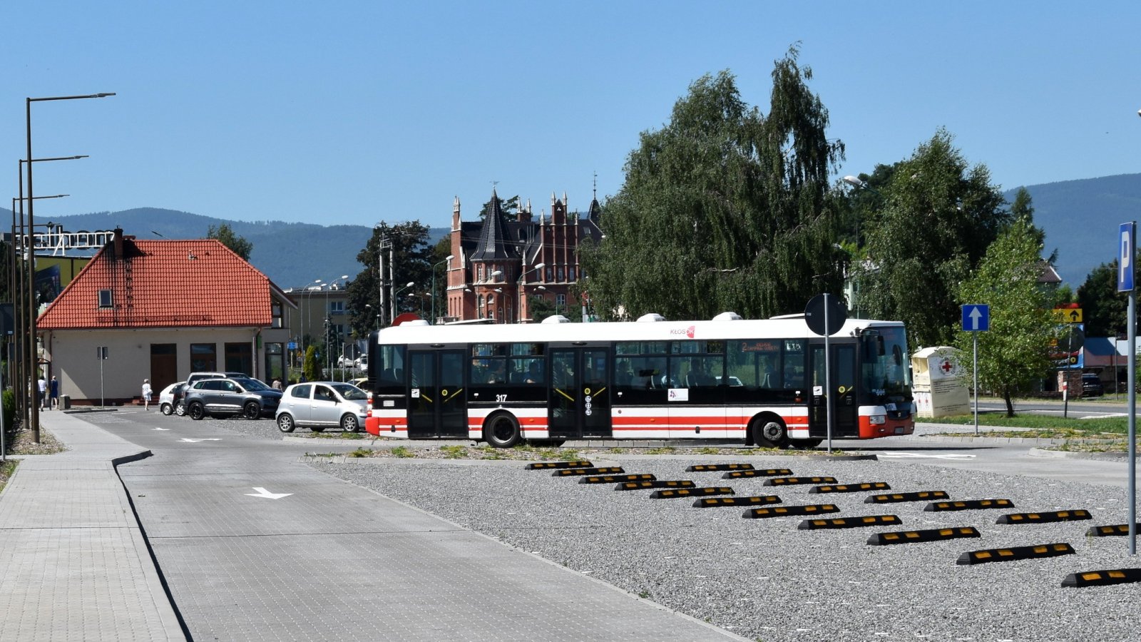 Autobus w białe i czerwone poziome pasy stojący na parkingu, za autobusem duże drzewo