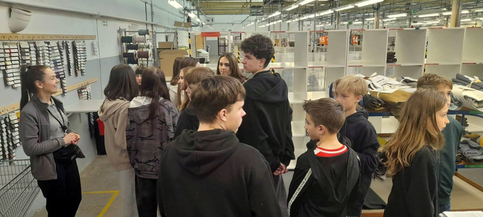 Kilkunastu uczniów w hali produkcyjnej, w tle takniny i nici