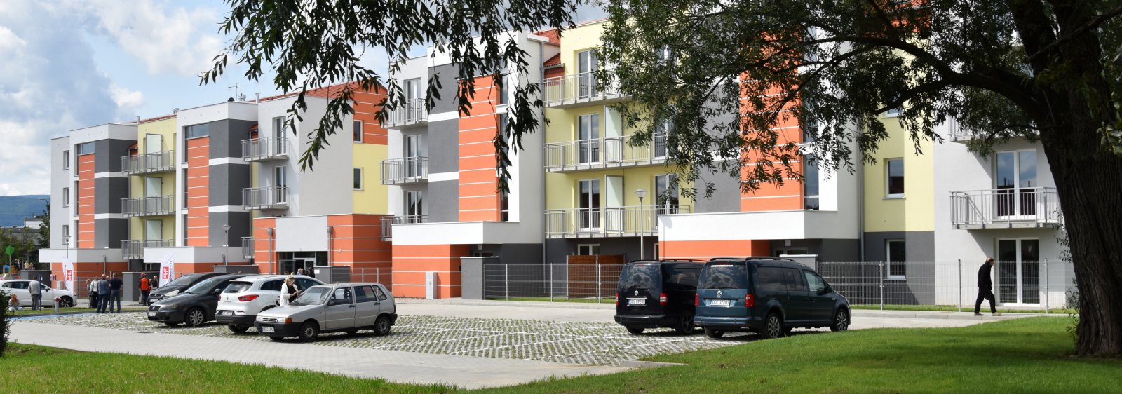 Nowy trzypiętrowy budynej z białą i pomarańczową elewacją, przed nim duży parking