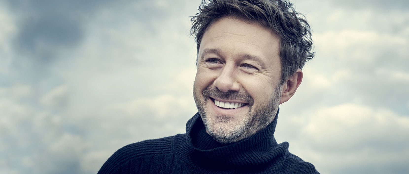 Portret uśmiechniętego artysty, w tle chmury. Fot: oficjalna strona artysty