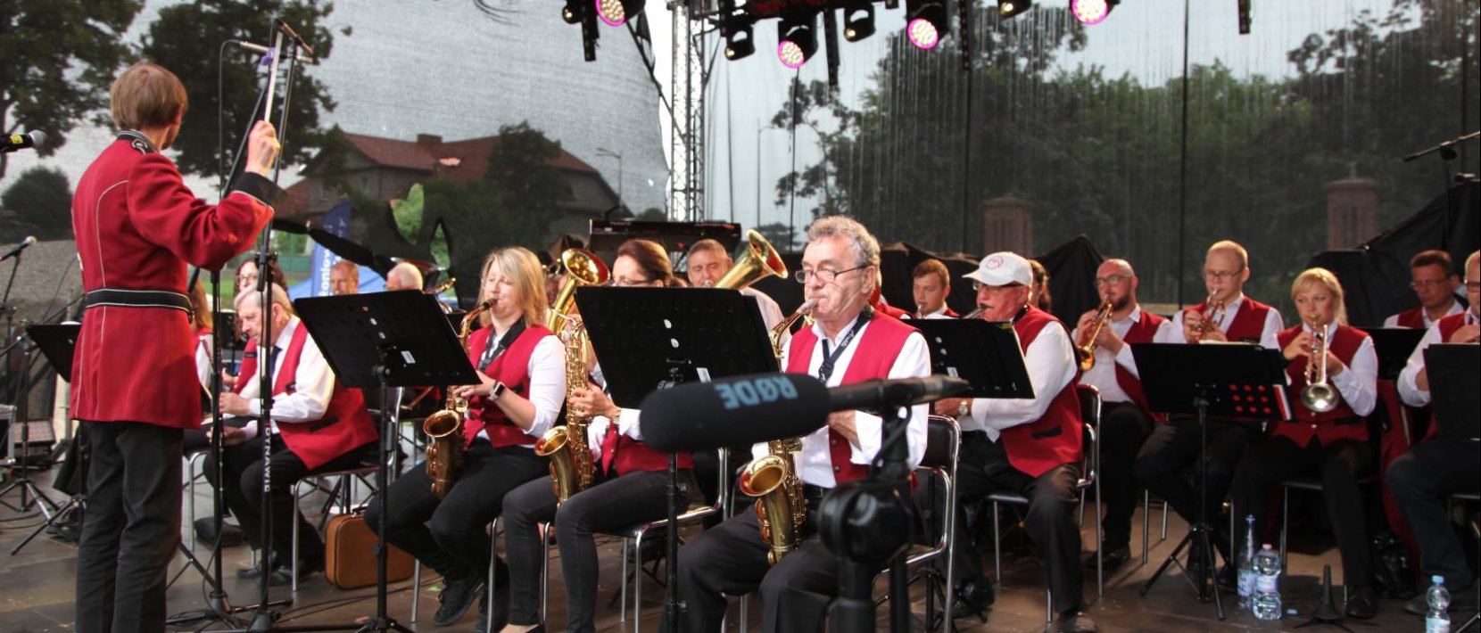 Koncert orkiestry dętej, muzycy w czerowno-białych strojach po lewej stronie dyrygent