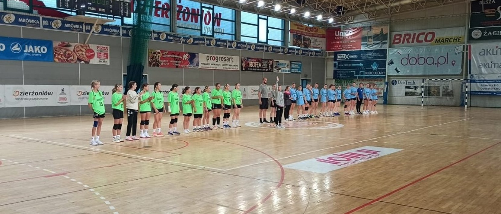 Dwie drużyny dziewczęce stojące równo na środku drewnianego parkietu w hali sportowej