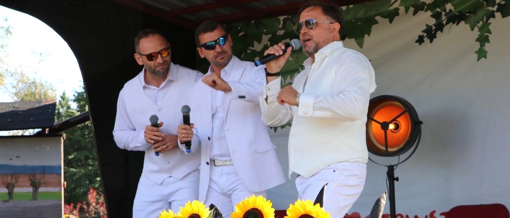 Trzech mężczyzn w białych strojach śpiewa na niewielkiej scenie