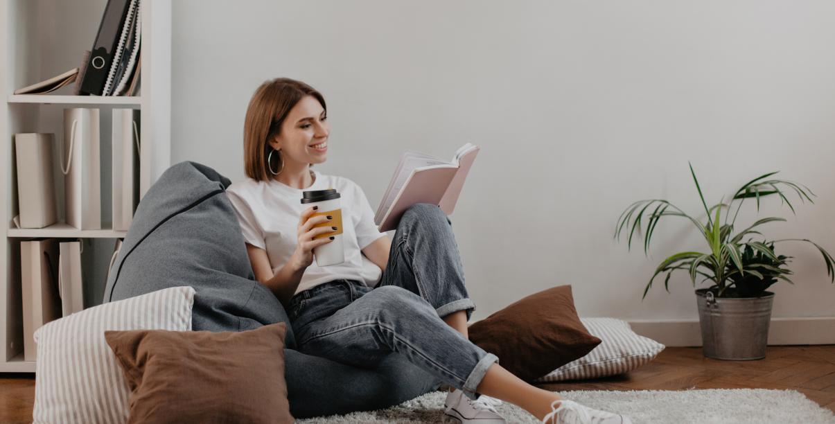 Kobieta siedząca na podłodze, wśród poduszek, czyta ksiązkę i pije herbatę