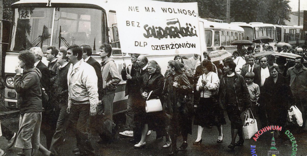 Czarno biała fotografia grupy ludzi przy autobusie z transparentem solidarności