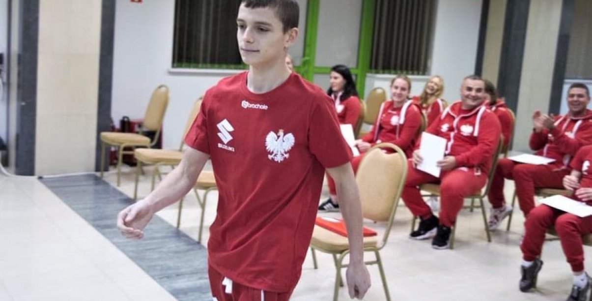 Młody zawodnik idący w koszulce z godłem, w tle gupa siedzących osób