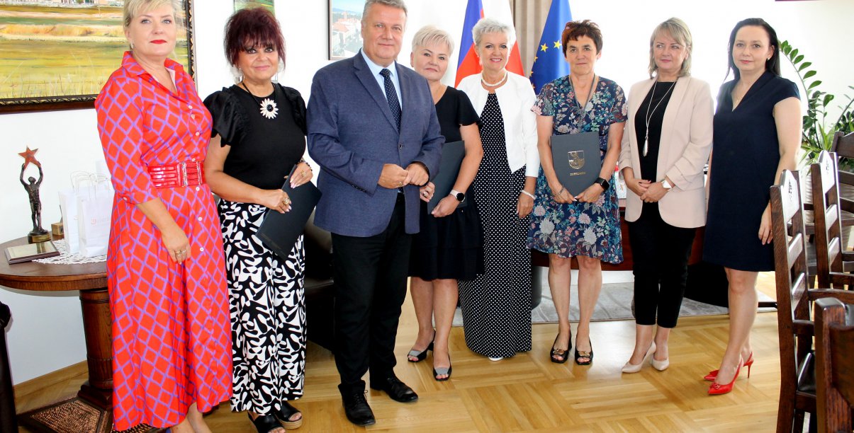 Siedem kobiet i mężczyzna stoją w jasnym pomieszczeniu, wszyscfy patrzą w obiektyw aparatu, w tle flagi