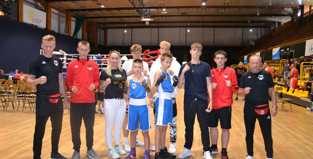 Grupa bokserów i bokserek w hali sportowej, po bokach trenerzy młodych zawodników