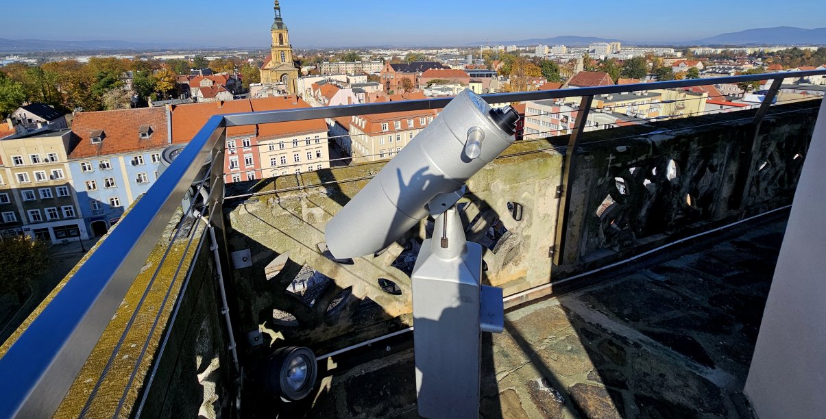 Wieża ratuszowa z kamiennymi i metalowymi barierkami, na pierwszym planie luneta, w tle panorama miasta