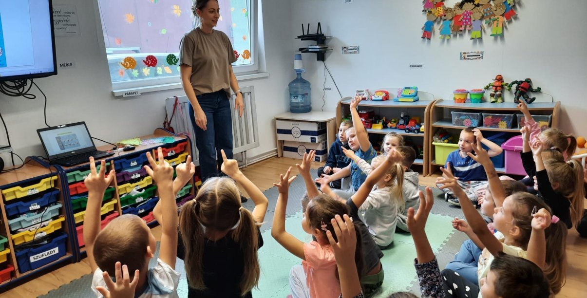 Przedszkolaki siedzący w grupie zgłaszają się do odpowiedzi, przed nimi młoda kobieta