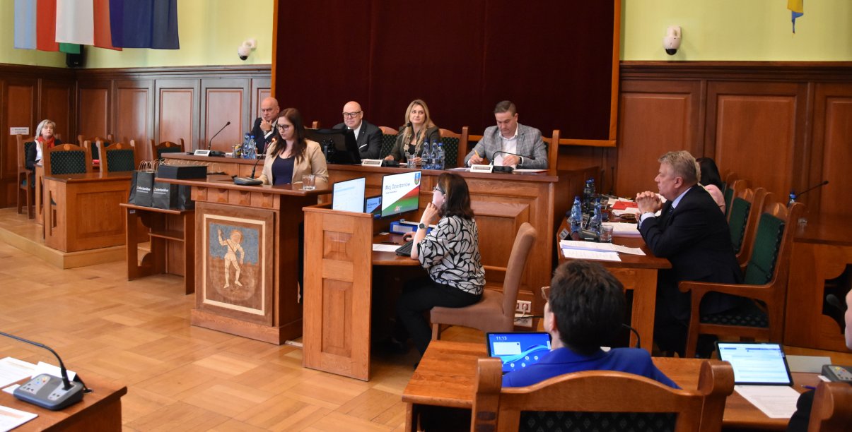 Sala obrad, za stopłem prezydialnym siedzą cztery sosoby, przed stołem mównica