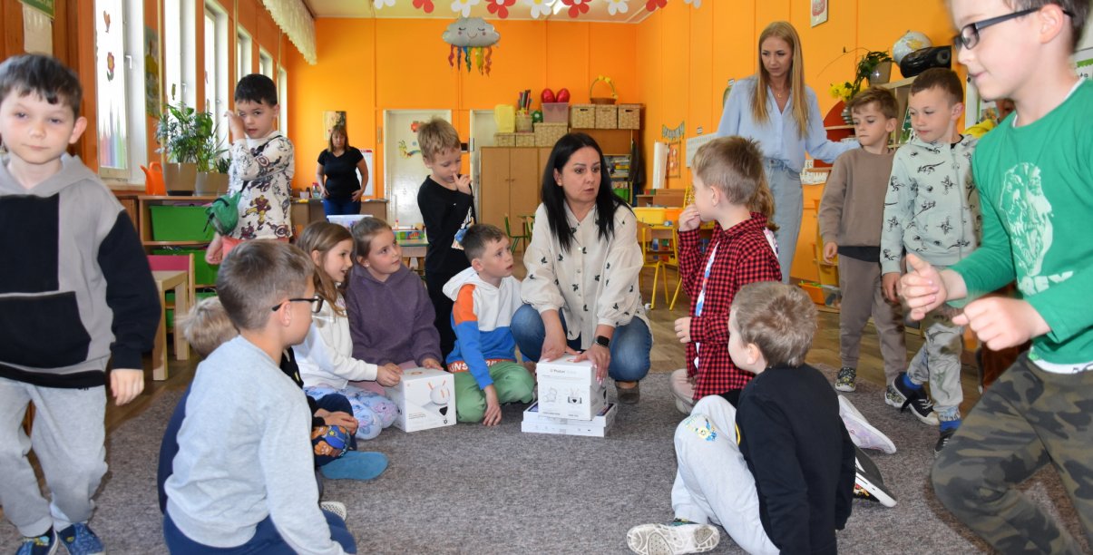 Grupa przedszkolaków w dużej sali, siedzących na podłodze i rozpakowujących z pudełek roboty i tablety