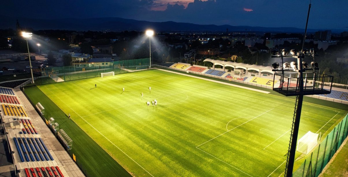 Stadion piłkarski nocą, oświetlony, z trybunami po bokach, na muraiwe grupka piłkarzy