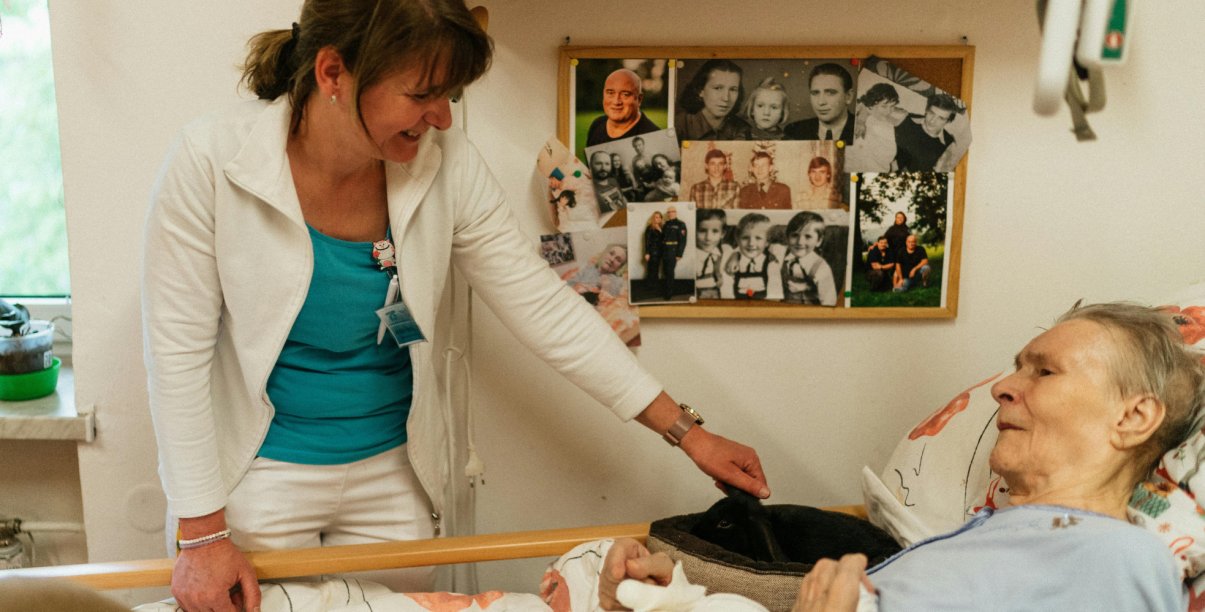 Starsza kobieta leżąca w łóżku, młodsza kobieta stojąca obok się nią opiekuje, na ścianie widoczne fotografie rodzinne
