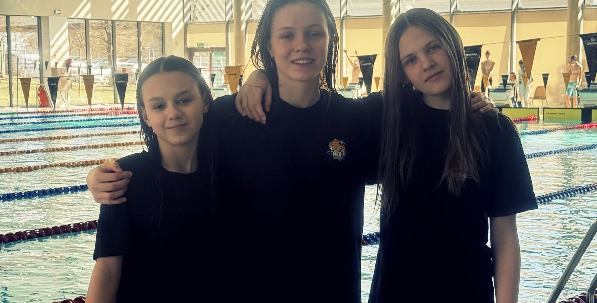 Trzy młode zawodniczki w czarnych koszulkach na tle basenu