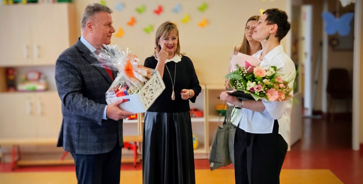 Burmistrz wręczający dyrektorce żłobka kwiaty i biały koszyk ze słodyczami