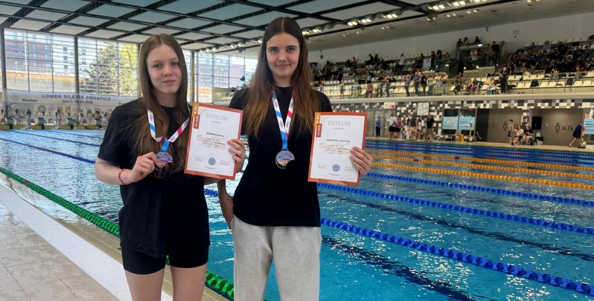 Dwie dziewczyny z medalami i dyplomami stoją przy krawędzi basenu