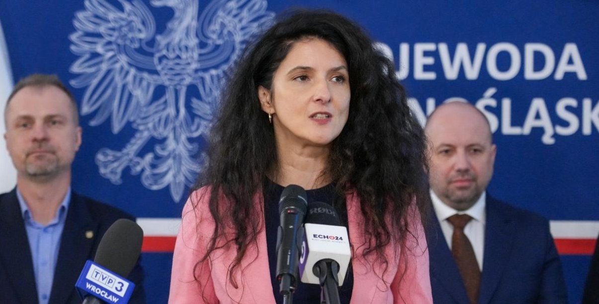 Wojewoda Dolnośląska przy mikrofonie, za nią dwóch mężczyzn na niebieskim tle