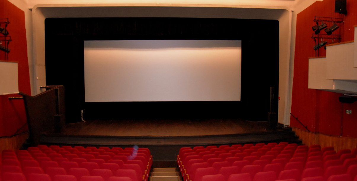 Biały ekran kinowy, przed ekranem scena i czerwone krzesełka, po bokach czerwone ściany