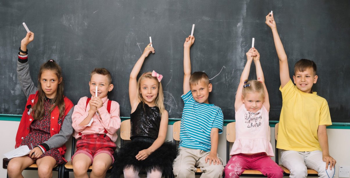 Trzech chłopców i trzy dziewczynki siedzą na krzesłach, za nimi tablica szkolna