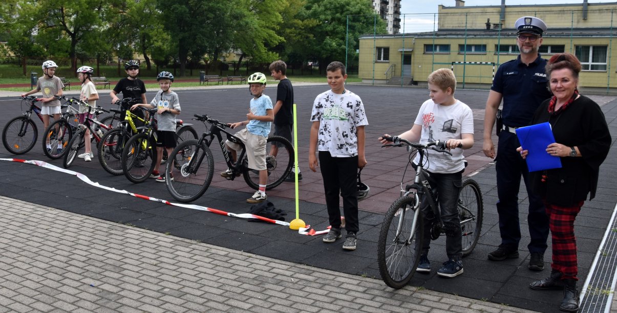 Grupa uczniów z rowerami ustawiona w szeregu, obok policjant i nauczycielka