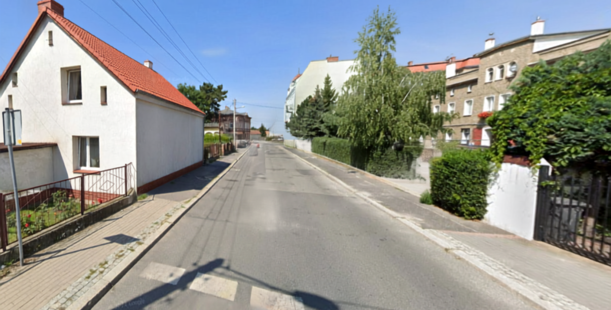 Droga asfaltowa, po lewej biały domek jednorodzinny, po praweej drzewa i domy jednorodzinne