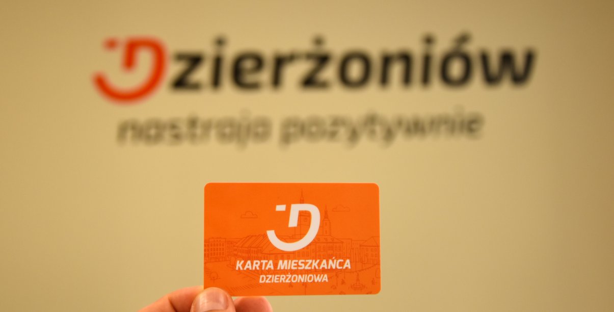 Dłoń trzymająca pomarańczową kartę mieszkańca na tle logotypu miasta