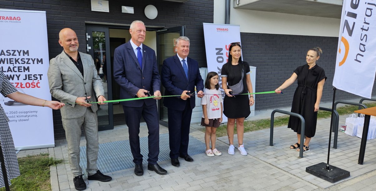 Trzech mężczyzn w garniturach i mama z dzieckiem przecinają zielona wstęgę przed wejściem do budynku