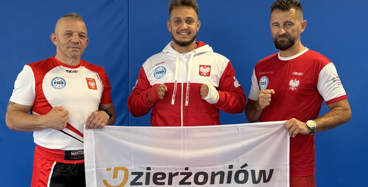 Maksymilian Pelej w biało-czerwonym dresie. Po bokach trenerzy trzymający flagę z logotypem Dzierżoniowa