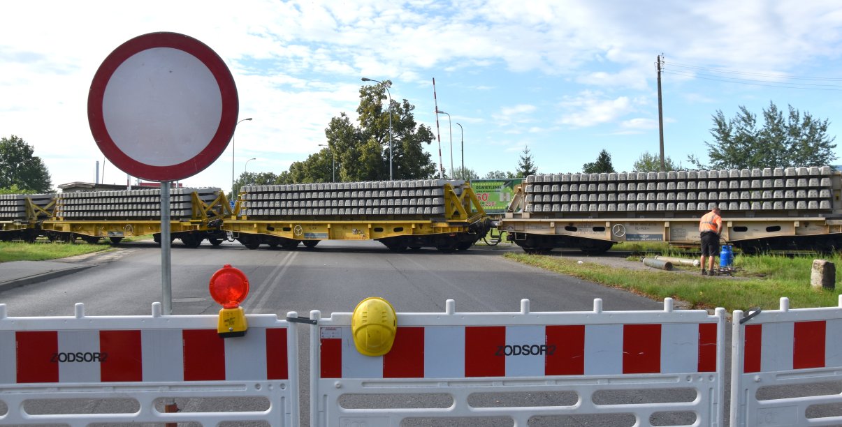 Przejazd kolejowy zagrodzony barierkami, na torach skład wiozący podkłady kolejowe