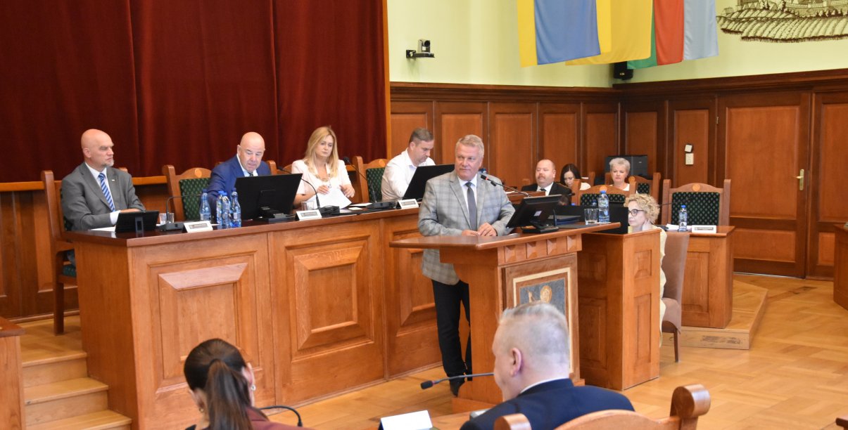 Sala obrad, przy mównicy burmistrz, za nim przewodnicząca i trójka wiceprzewodniczących przy stole prezydialnym
