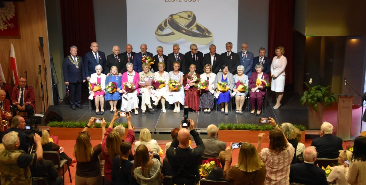 Jubilaci na wspólnym zdjęciu, kobiety siedzą na krzesłach, za nimi stoją mężowie. Przed sceną grupa osób robiących zdjęcia