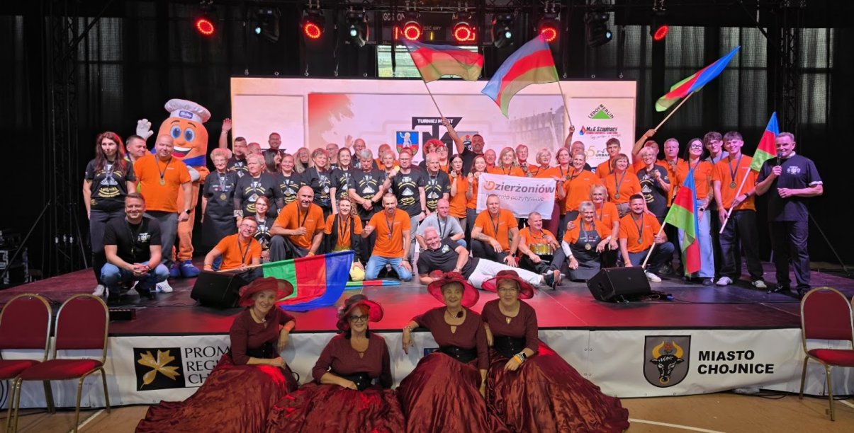 duża grupa osób na scenia w czarnych i pomarańczowych koszulkach, na pierwszym planie cztery panie w bordowych sukniach i kapeluszacj