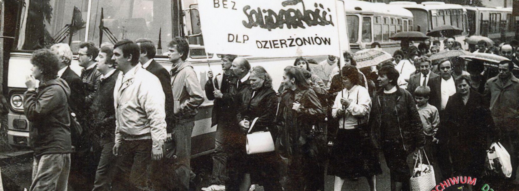 Czarno biała fotografia grupy ludzi przy autobusie z transparentem solidarności