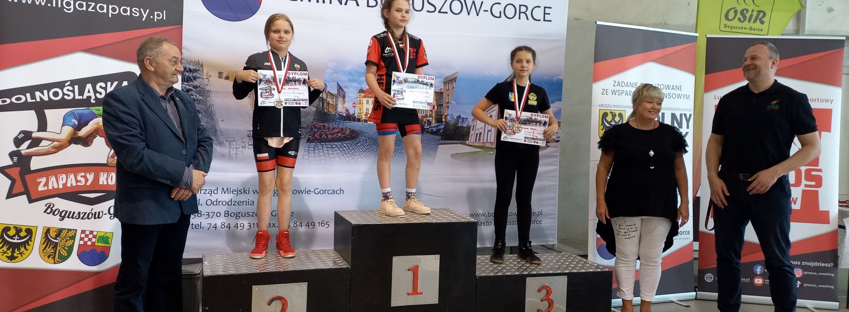 Trzy zapaśniczki na podium, obok podium trenerzy