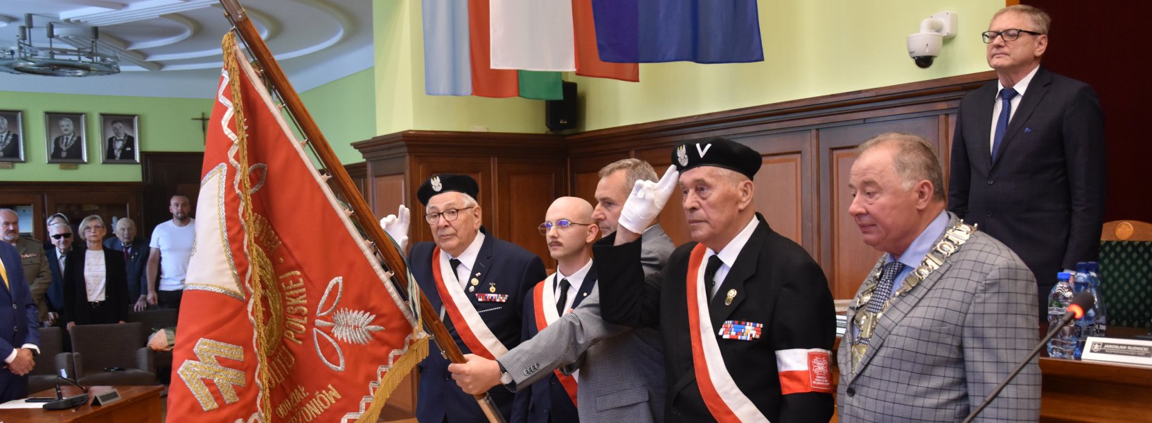 Kombatanci w mundurach trzymają czerwony sztandar i salutują