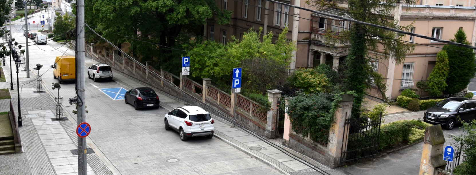 Dorga z kostki kamiennej widziana z góry, w tle zasłanięta drzewami i zielenią kamienica