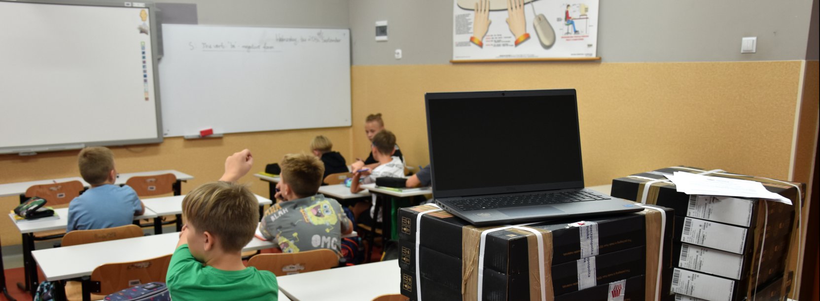 Uczniowie podczas zajęć w klasie, na pierwszym planie laptop i uczeń z podniesioną ręką 