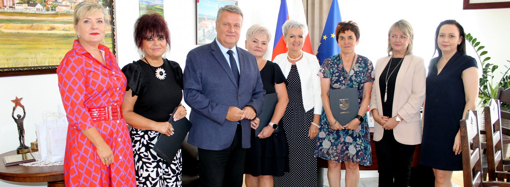 Siedem kobiet i mężczyzna stoją w jasnym pomieszczeniu, wszyscfy patrzą w obiektyw aparatu, w tle flagi