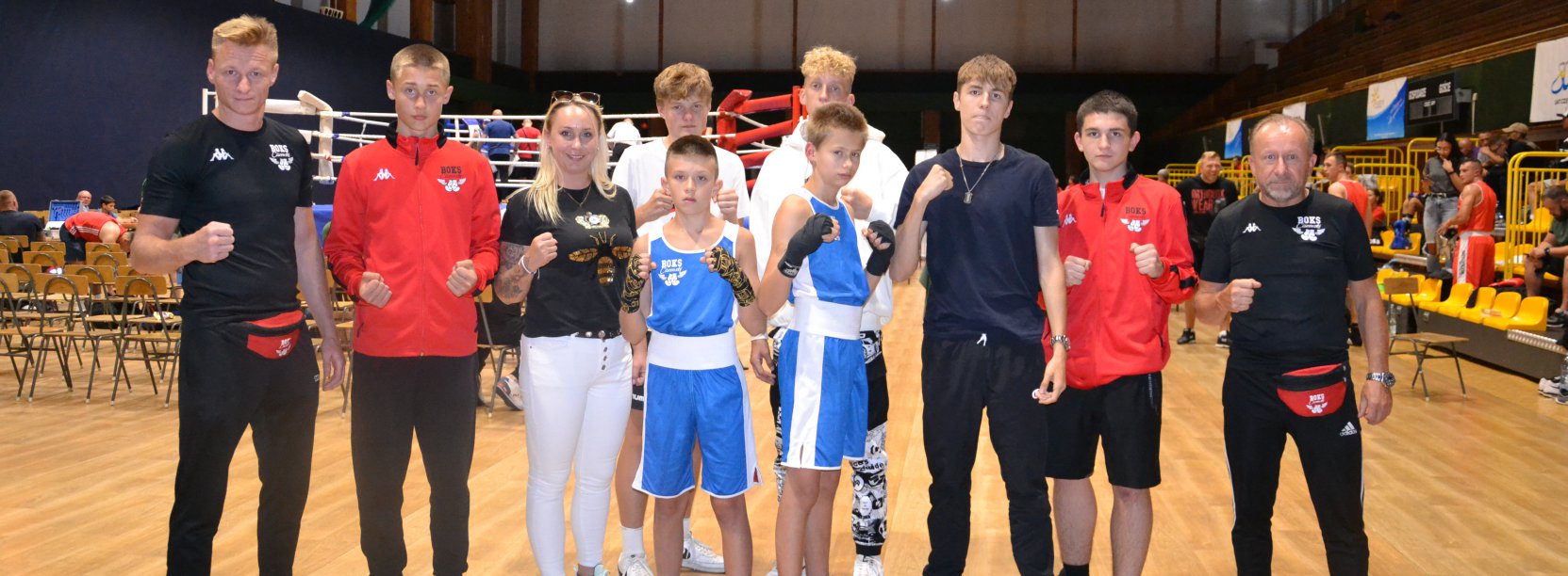 Grupa bokserów i bokserek w hali sportowej, po bokach trenerzy młodych zawodników